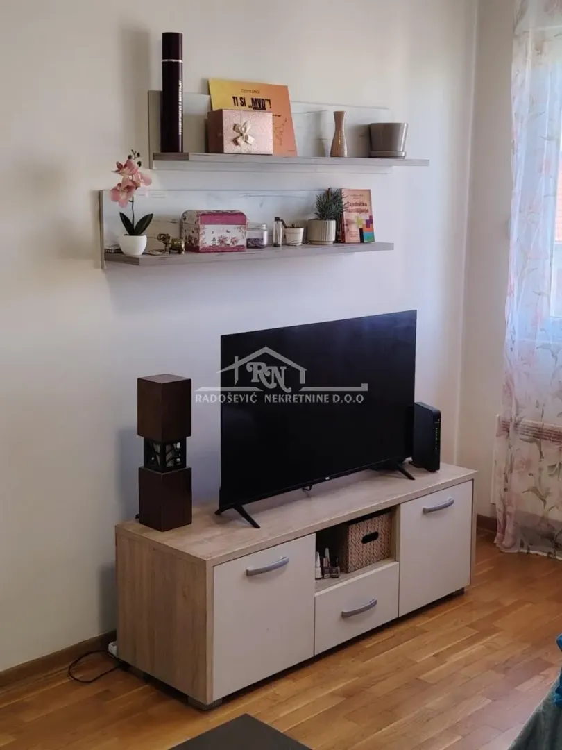 Dnevna soba sa TV komodom i policama na zidu.