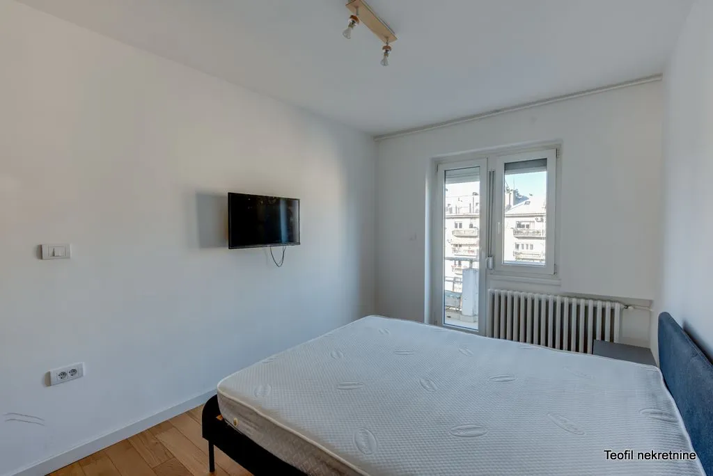 Spavaća soba sa bračnim krevetom, televizorom na zidu i izlazom na balkon.