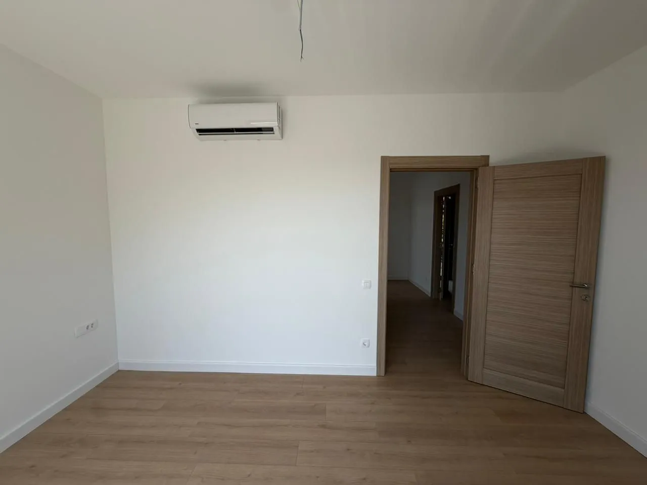 Prazna soba sa bijelim zidovima, drvenim podom i klima uređajem.