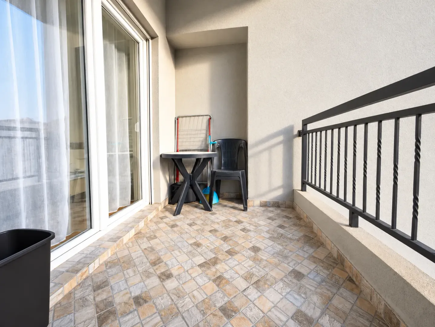 Nenamešten balkon sa keramičkim pločicama, stolom, stolicom i metalnom ogradom.