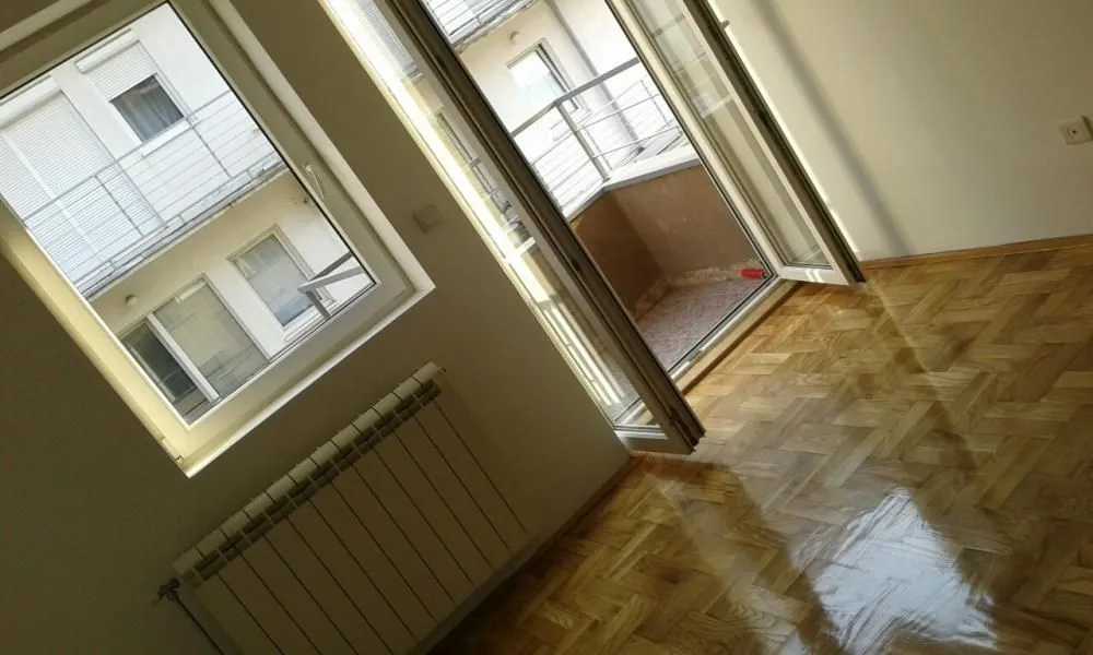 Prazna soba sa parketom, prozorom, radijatorom i izlazom na terasu.
