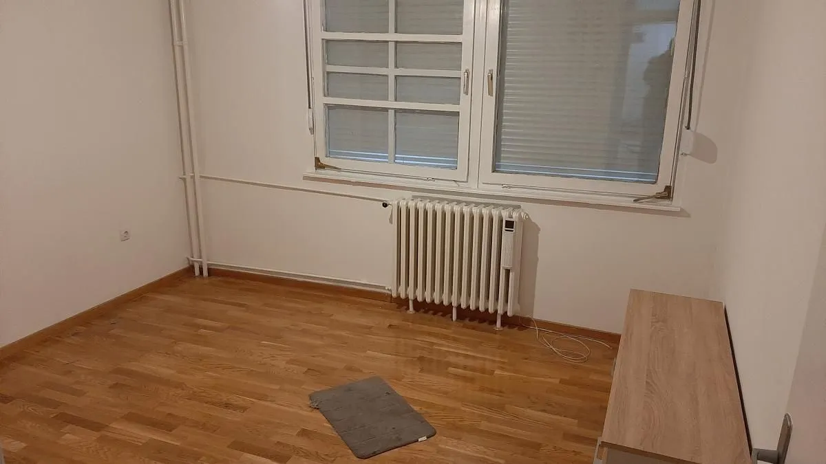 Prazna soba sa prozorom, drvenim podom i radijatorom.
