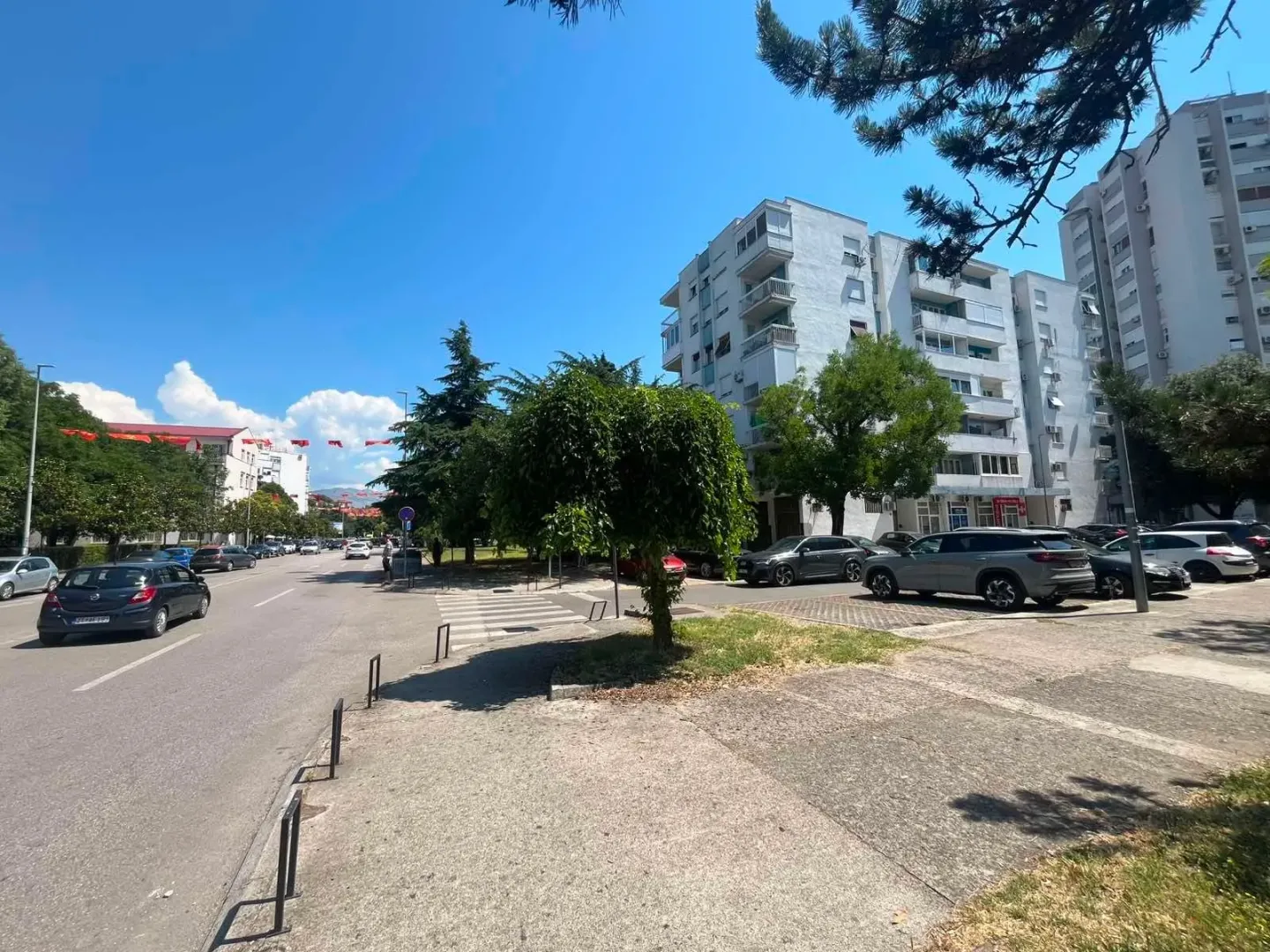 Pogled na ulicu ispred zgrade sa parkingom i drvećem.