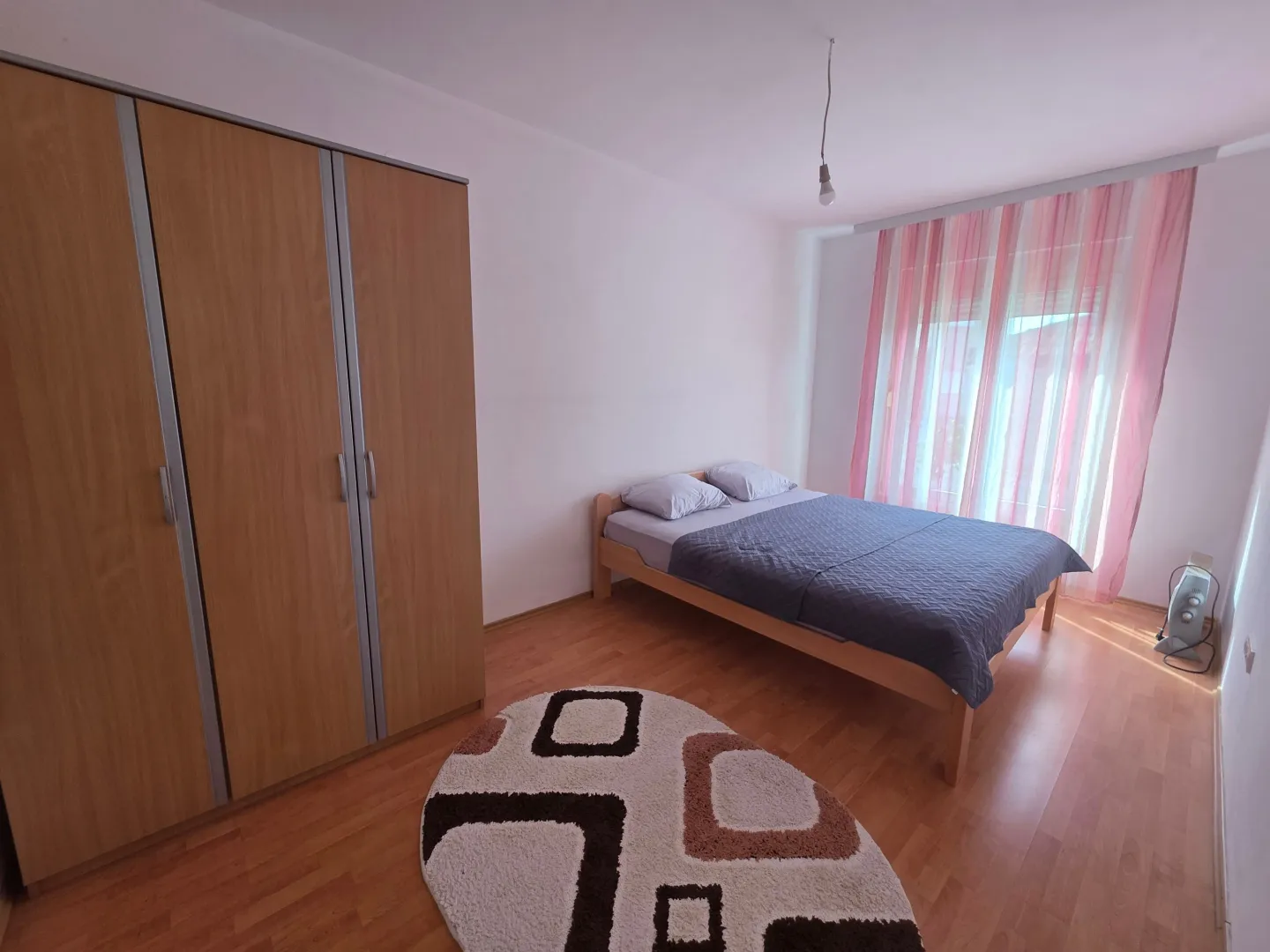 Furnished bedroom with a double bed, large wooden wardrobe, and a window with pink curtains.