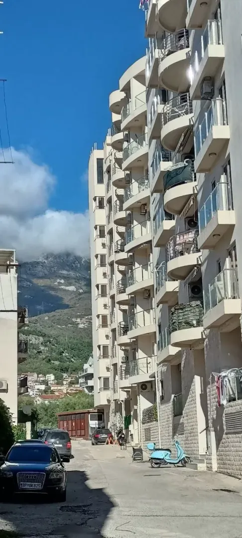 Pogled na spoljašnjost zgrade sa terasama i parkiranim automobilima ispred.