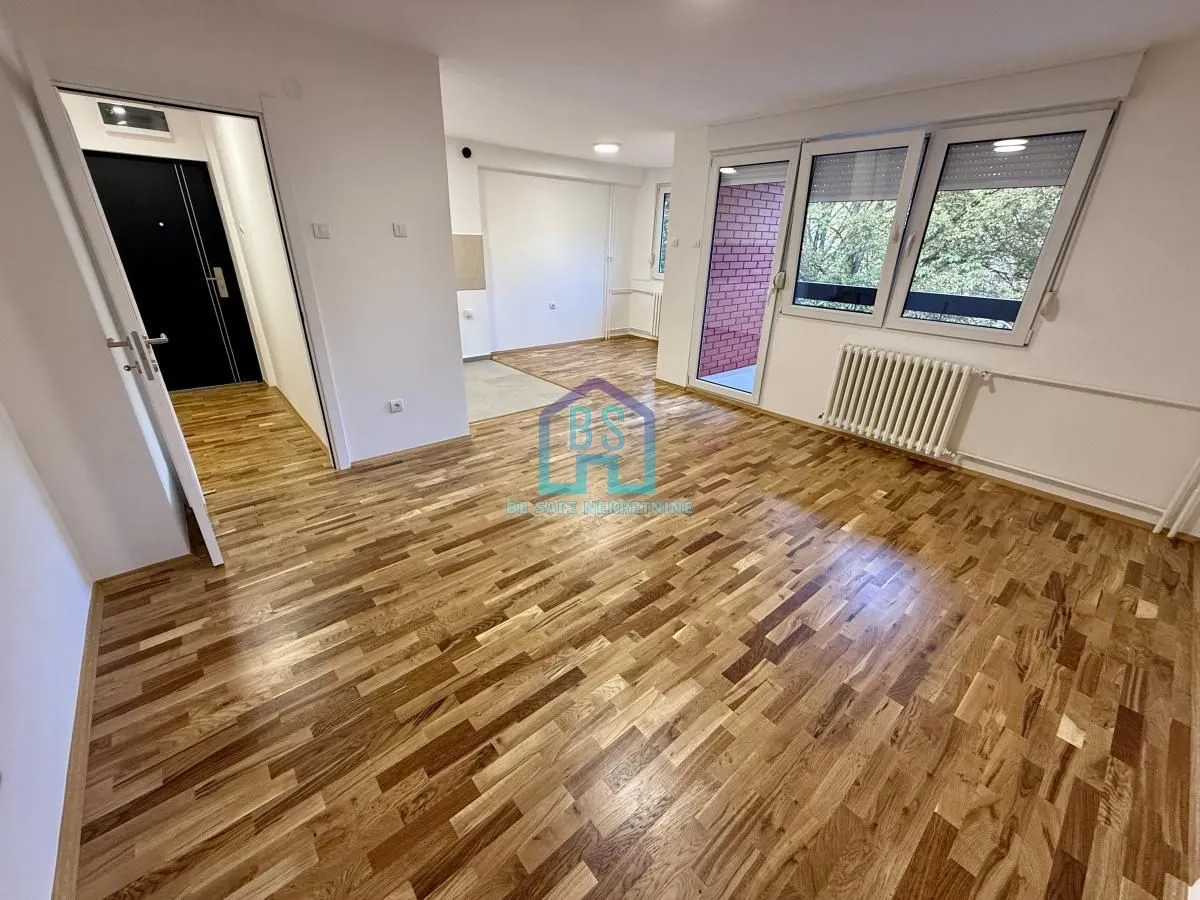 Prazna dnevna soba sa drvenim parketom, prozorima i vratima koja vode na balkon.