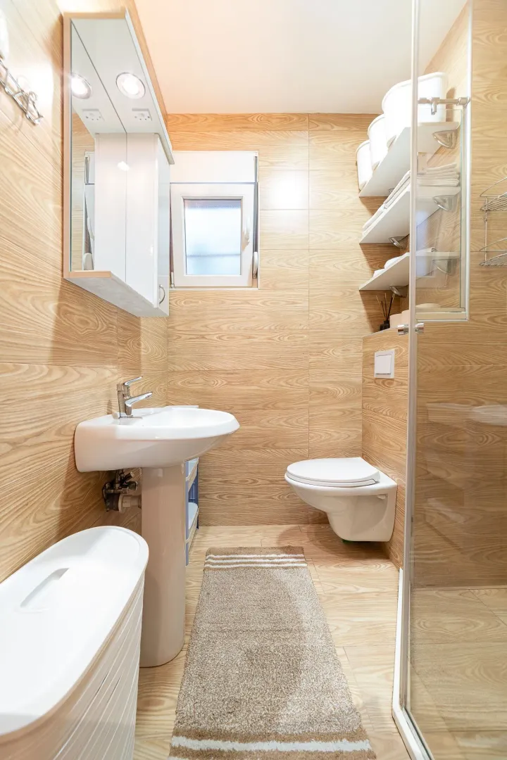 Bathroom lined with wood-textured ceramic tiles, featuring a sink, toilet, and shower cabin.
