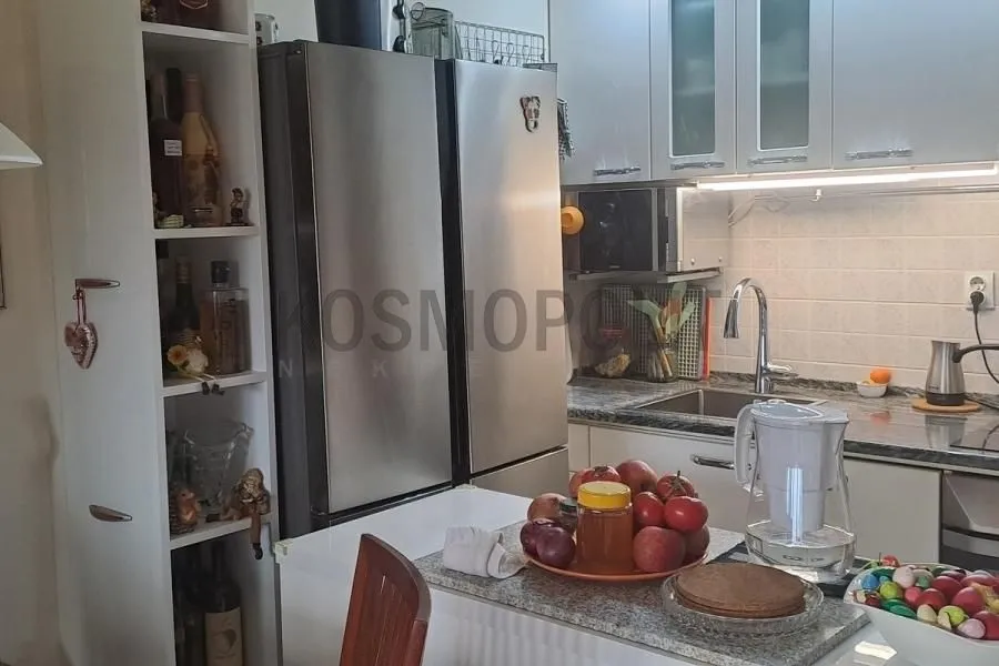 Manja kuhinja sa belim elementima, frižiderom i radnom površinom.