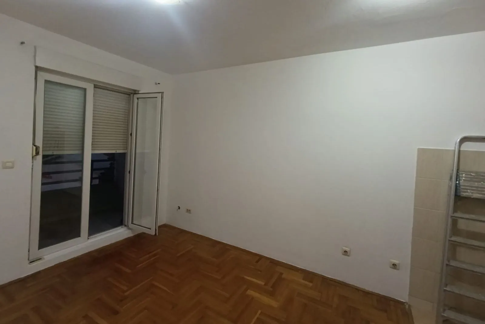 Prazna prostorija sa bijelim zidovima, drvenim parketom i balkonskim vratima.