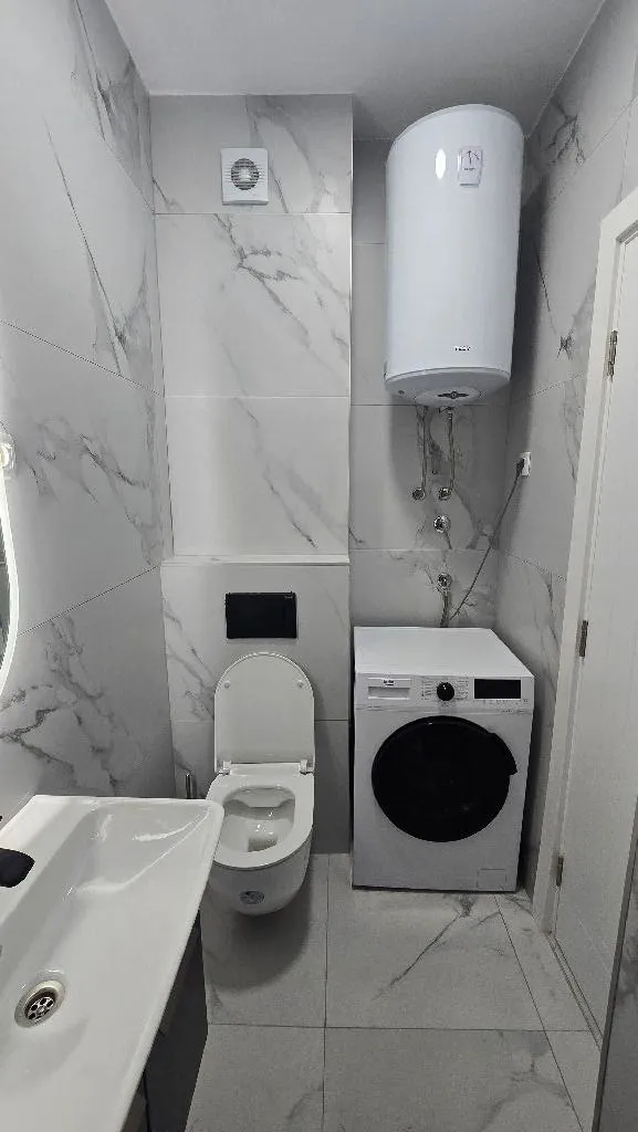 Modern bathroom with white tiles, toilet, washbasin, washing machine, and water heater.