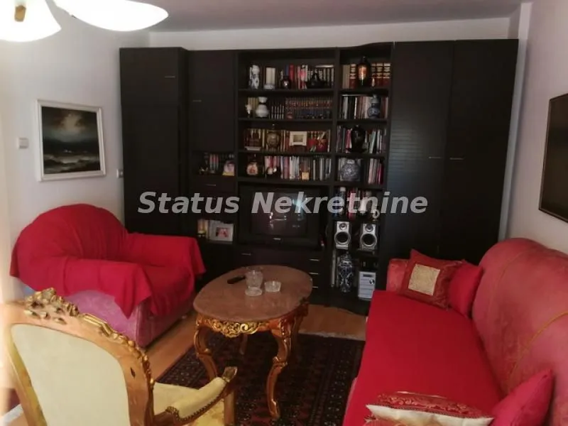 Dnevna soba sa garniturom, foteljom i velikim zidnim ormanom sa policama za knjige.