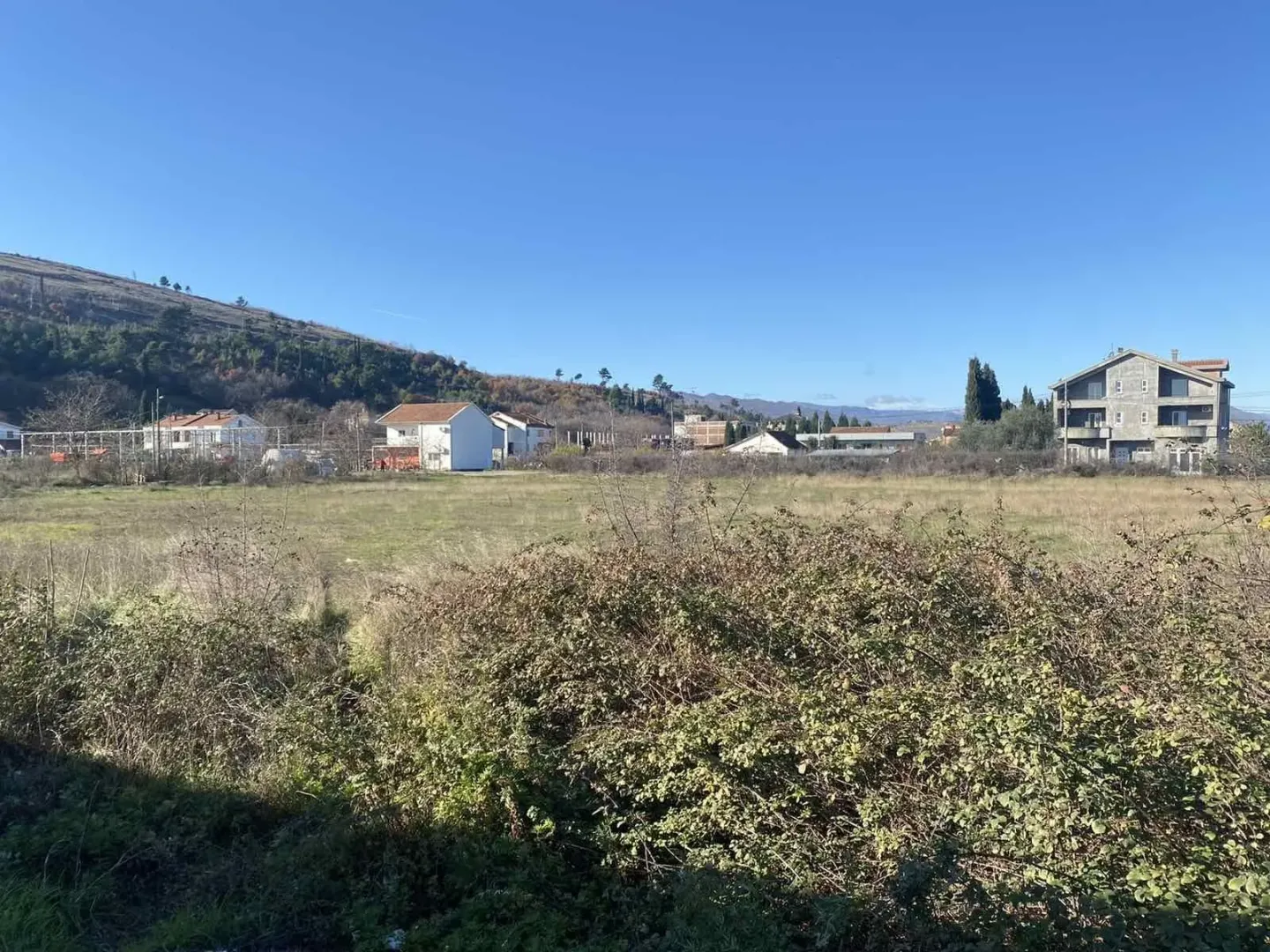 Prostrani ravni plac okružen kućama i zelenilom pod plavim nebom.