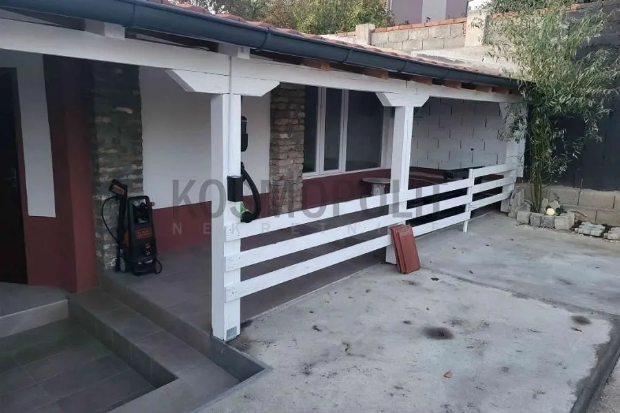 Covered patio in front of the house with a white wooden fence and tiled flooring.