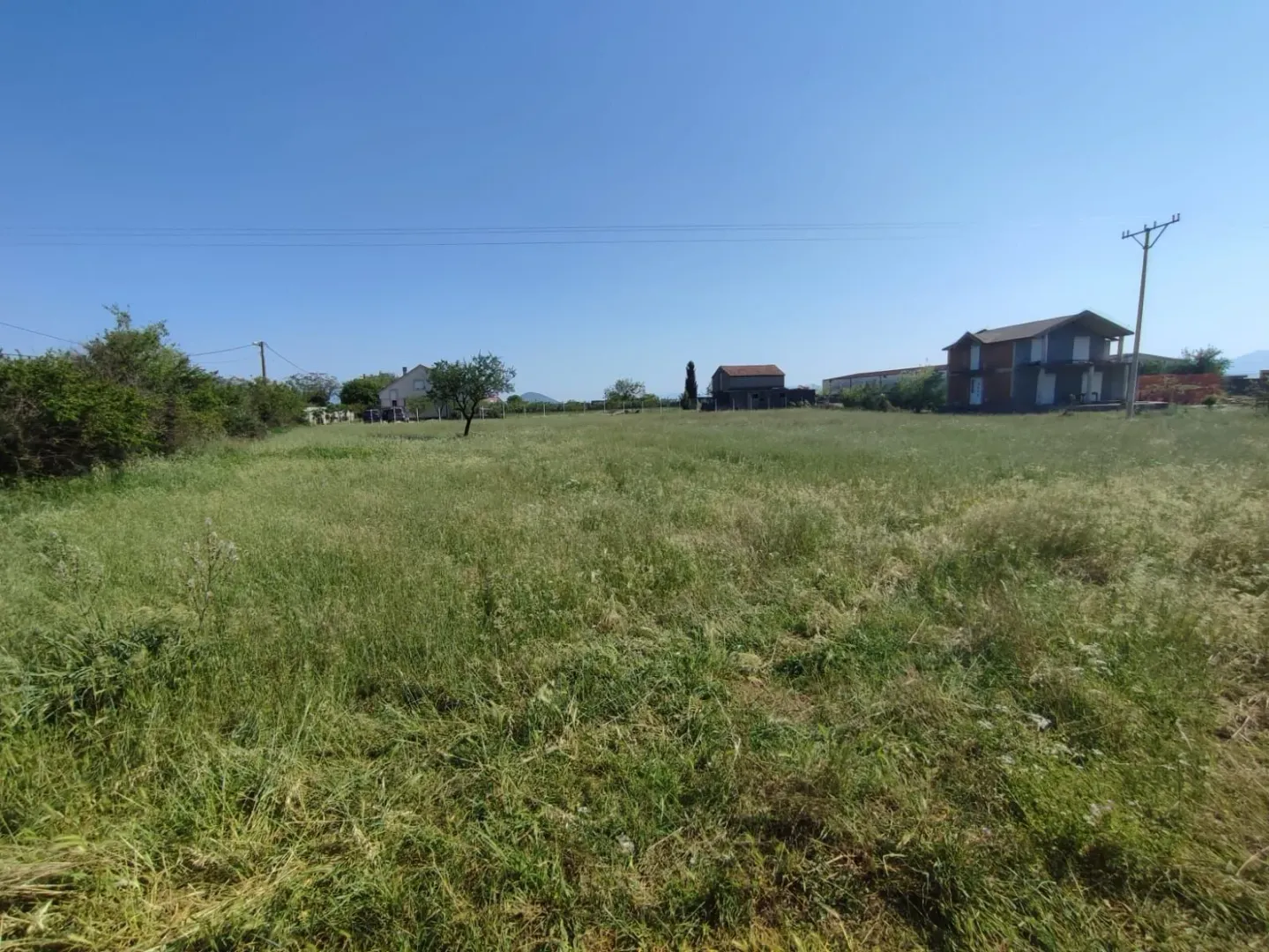 Prostran travnati plac sa kućama u pozadini pod vedrim nebom.