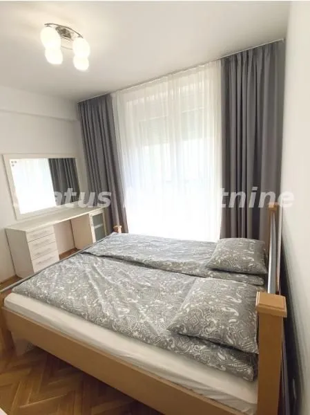 Nameštena spavaća soba sa bračnim krevetom, belom komodom i zavesama.