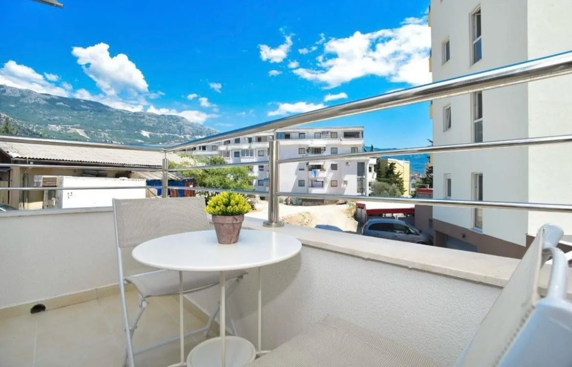 Bright balcony with a table, chairs, and a view of surrounding buildings and mountains.