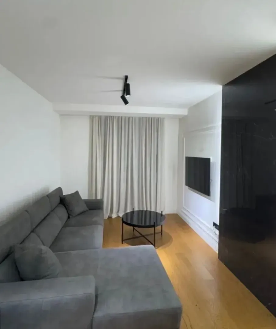 Moderan dnevni boravak sa ugaonom garniturom, stočićem i televizorom na zidu.