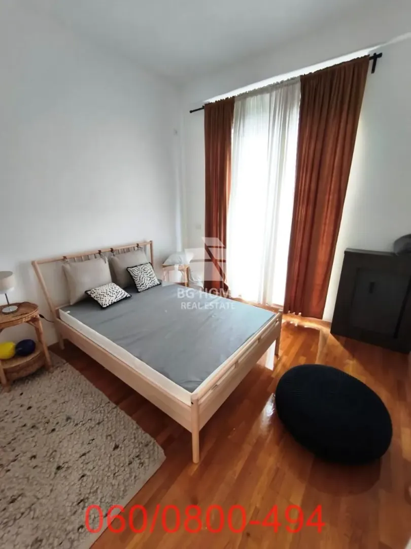 Nameštena spavaća soba sa bračnim krevetom, drvenim podom i prozorom sa zavesama.