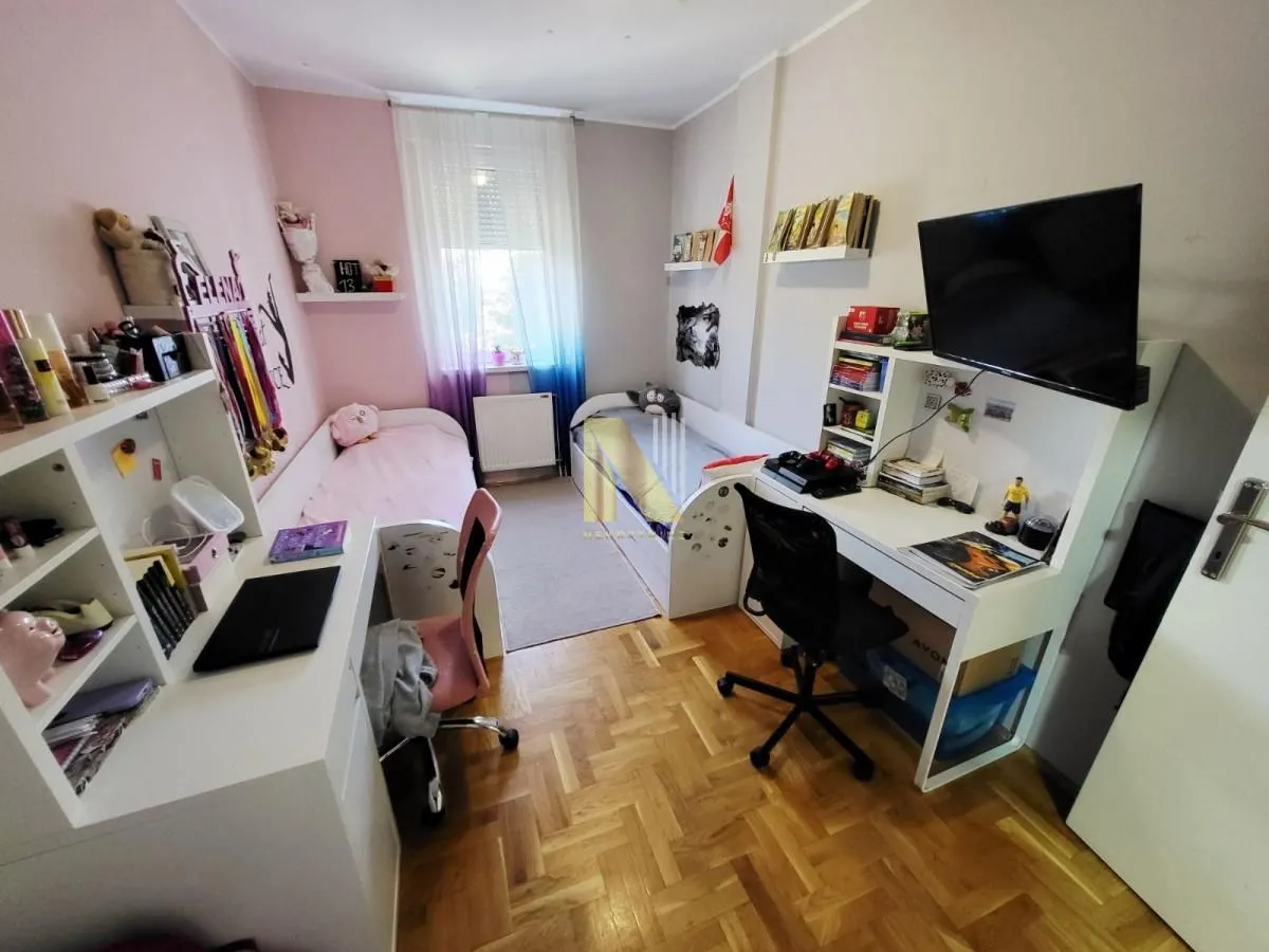 Dečja soba sa dva kreveta, radnim stolovima i televizorom.