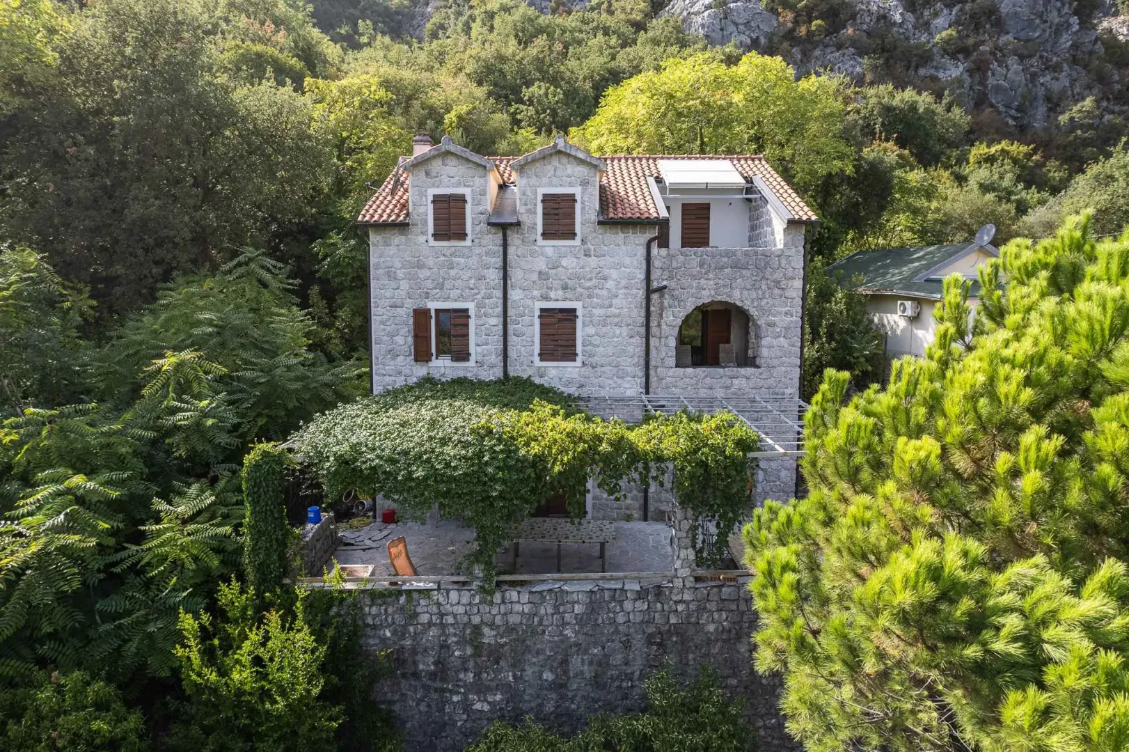 Pogled sa prednje strane na kamenu kuću sa terasom i zelenilom.