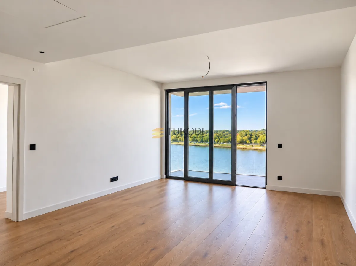 Empty bright room with wooden flooring and a river view through glass doors.
