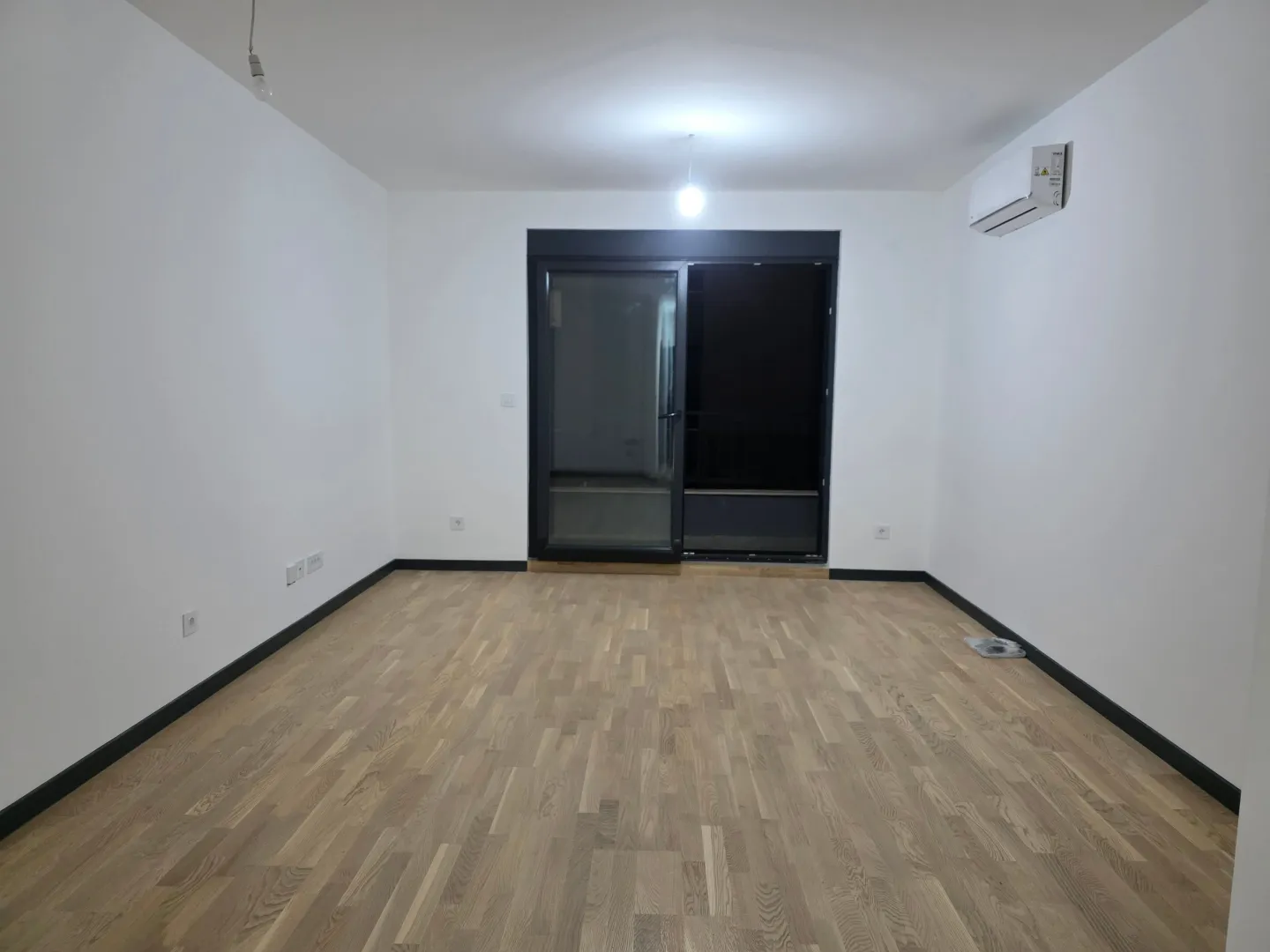 Prazna soba sa parketom, bijelim zidovima i klima uređajem.