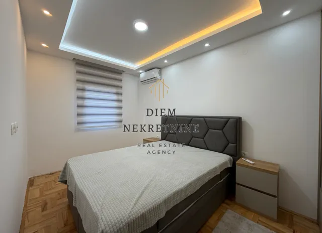 Furnished bedroom with a double bed, parquet flooring, and a dropped ceiling with lighting.