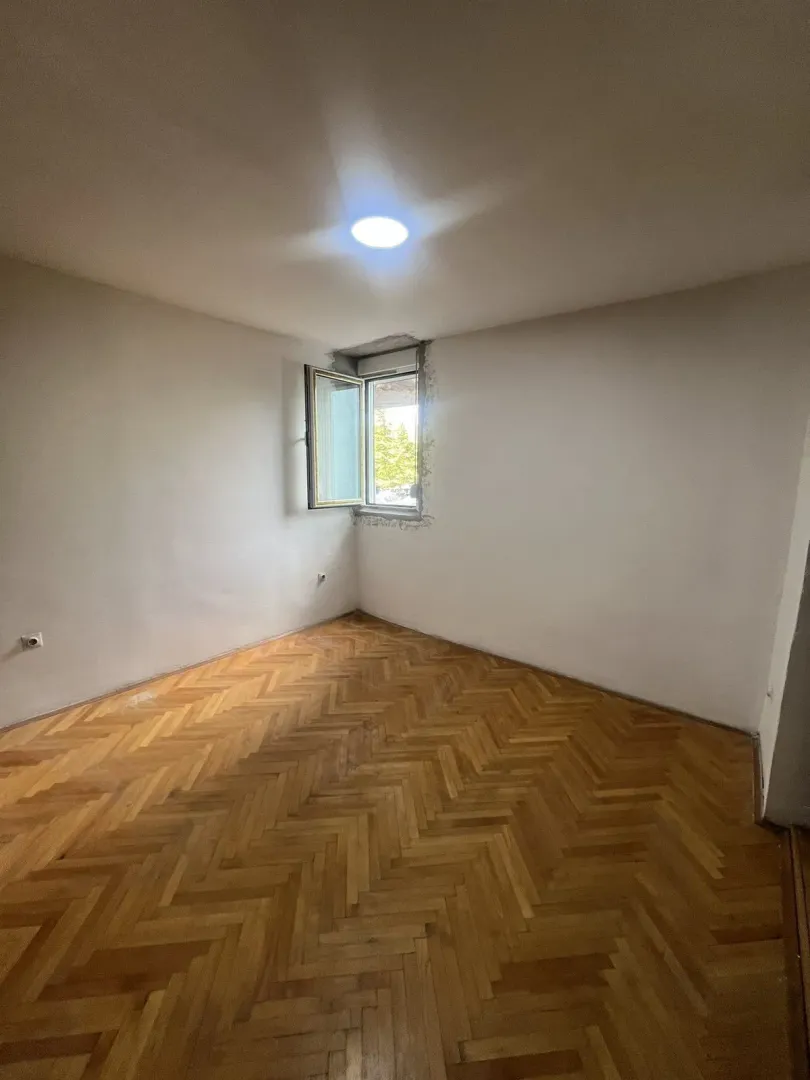 Prazna soba sa parketom i jednim otvorenim prozorom.