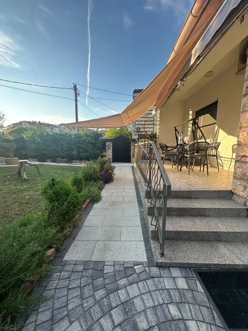 Dvorište kuće sa popločanom stazom, terasom i zelenilom.