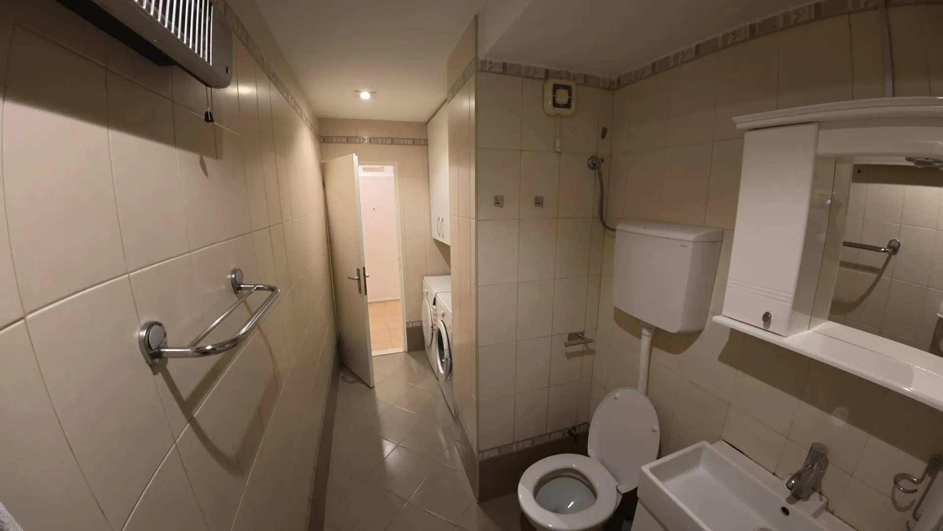 Smaller bathroom tiled in light colors with a toilet, washbasin, and washing machine.