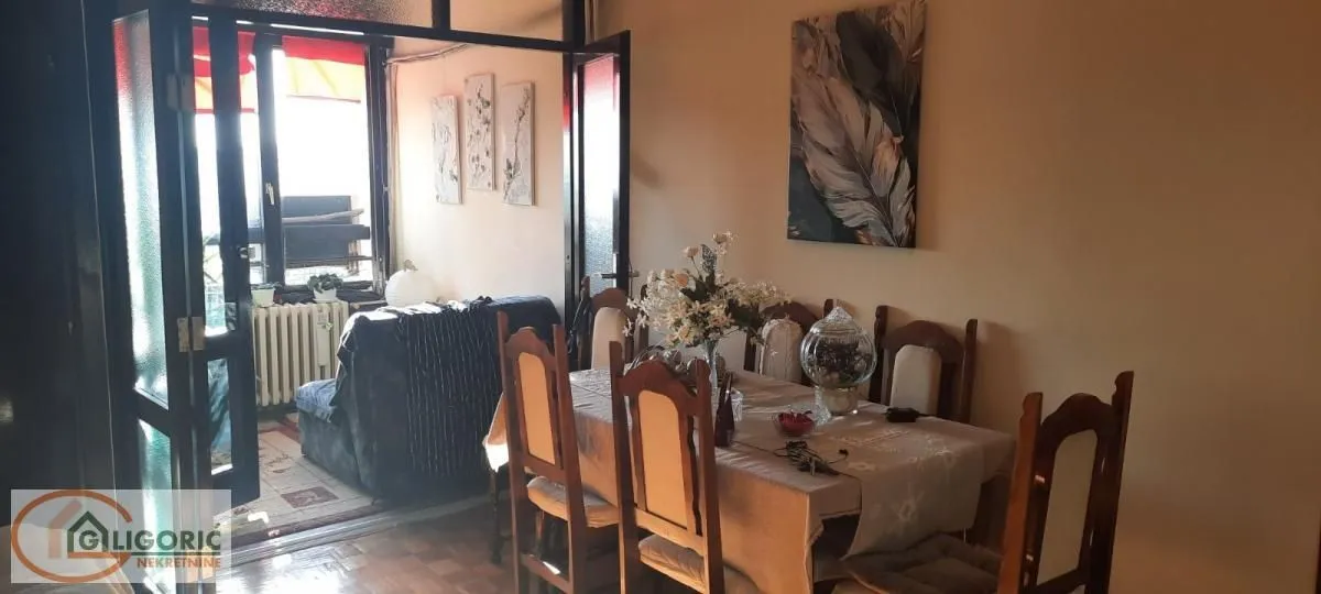 Trpezarija sa drvenim stolom i stolicama, sa pogledom na zastakljenu terasu.