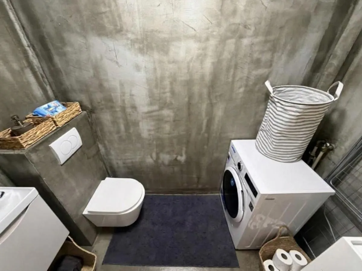 Kupatilo sa betonskim zidovima, wc šoljom, umivaonikom i mašinom za veš.