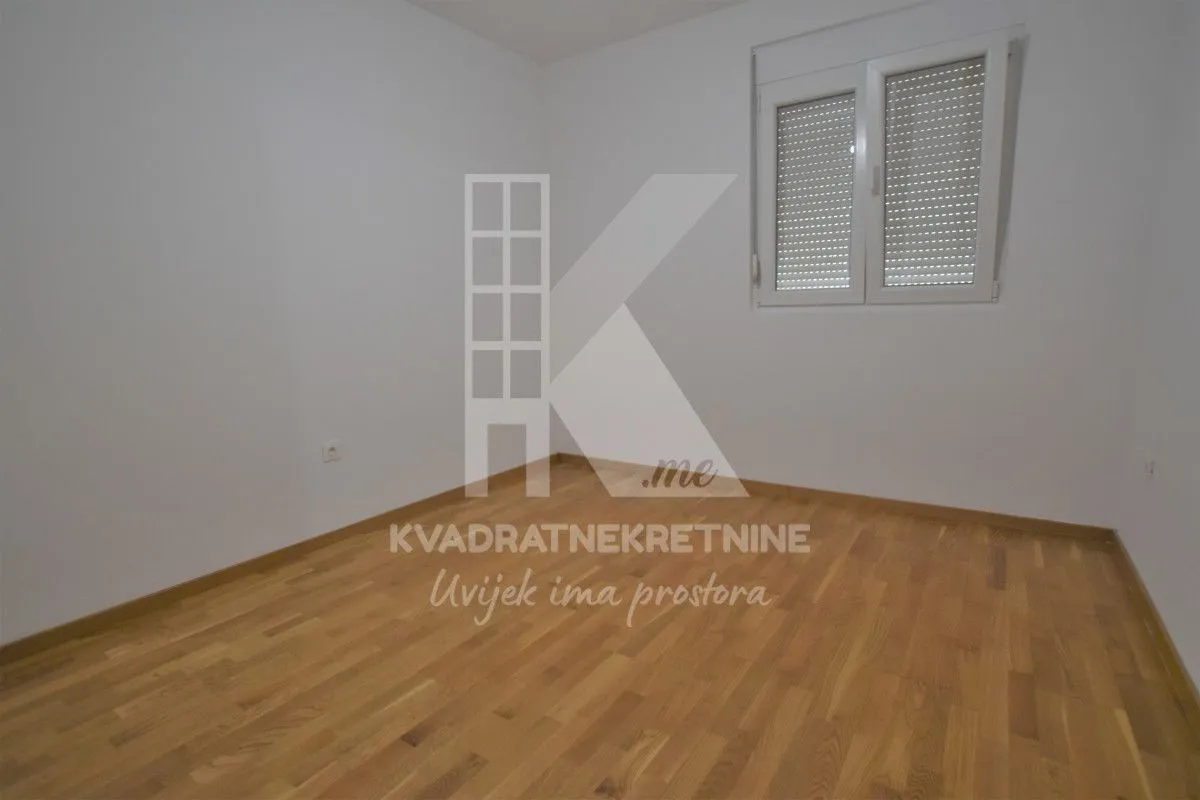 Prazna soba sa bijelim zidovima, parketom i prozorom sa roletnama.