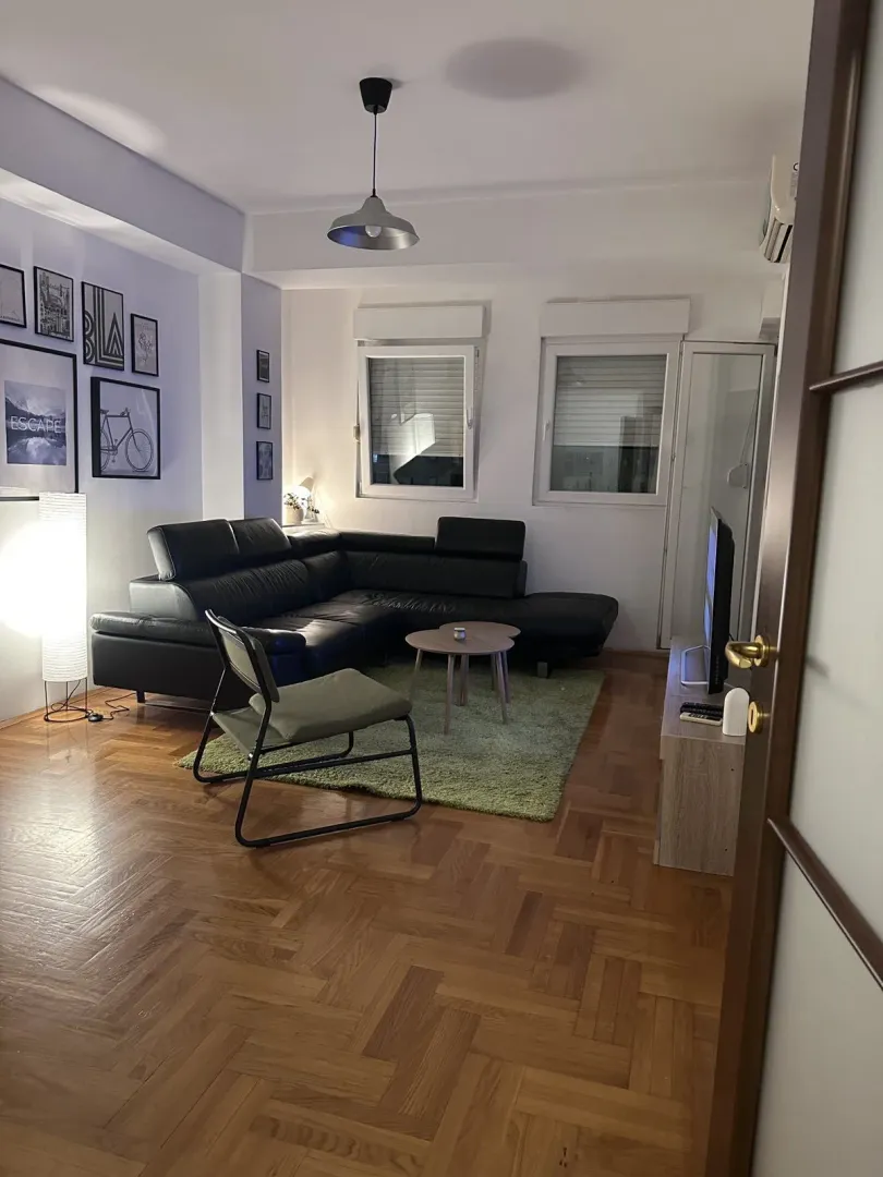 Bright living room with a black leather sectional sofa, coffee table, and wooden parquet floor.