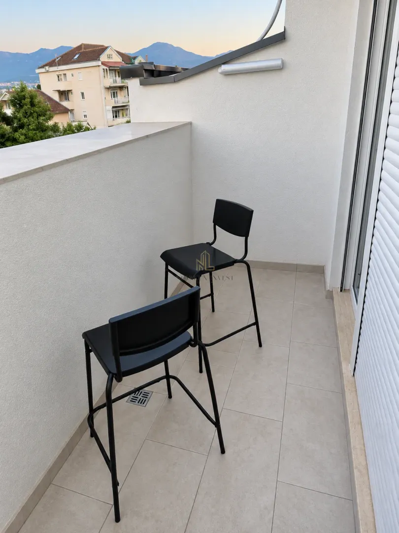 A small tiled balcony with two black bar stools and a view of surrounding buildings and hills.