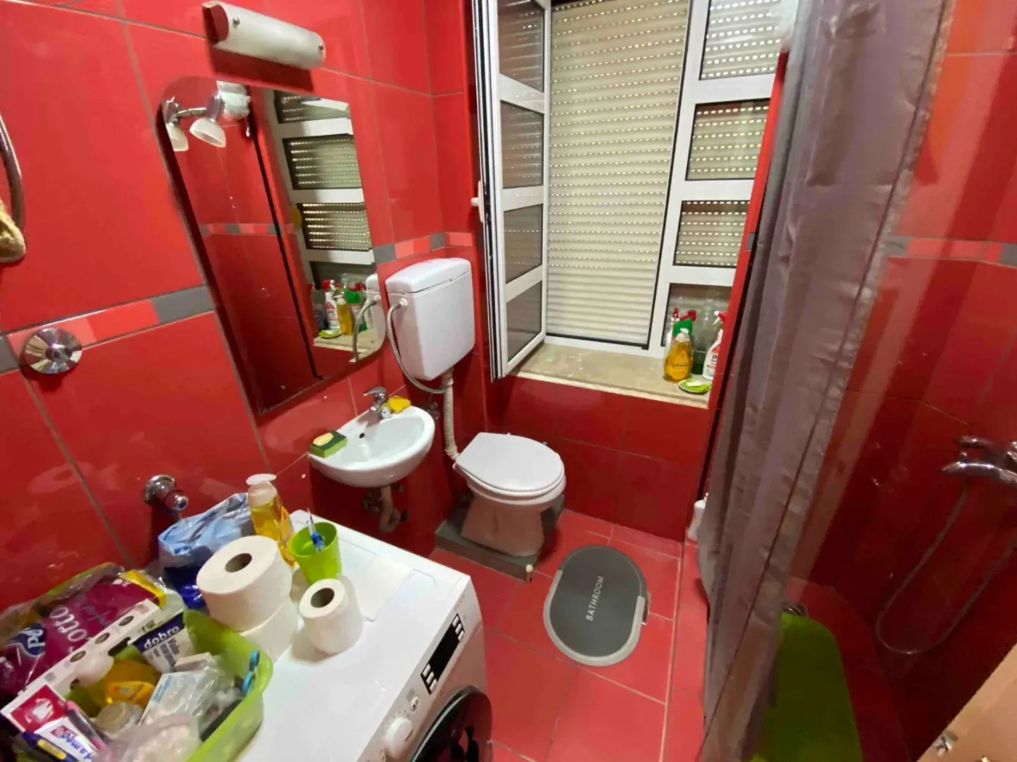 Manje kupatilo sa crvenim pločicama, umivaonikom, WC šoljom i veš mašinom.