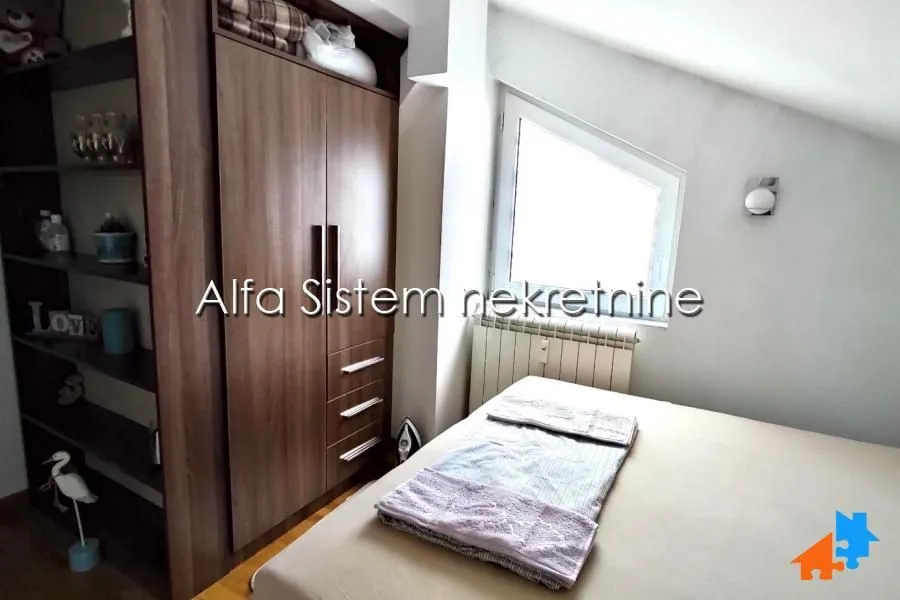 Nameštena spavaća soba sa bračnim krevetom i drvenim ormarom pod kosim krovom.