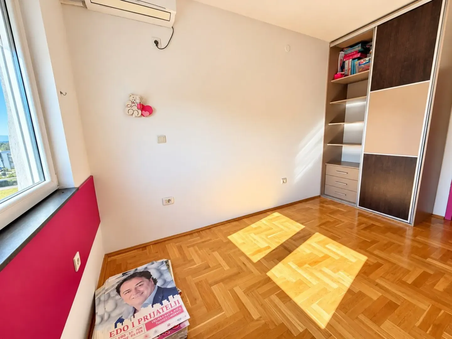 Prazna soba sa parketom, ugradnim ormarom i prozorom.
