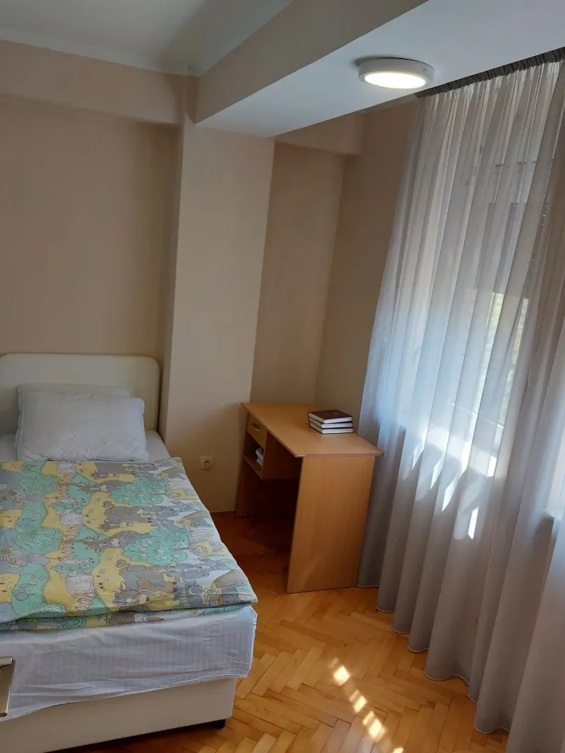 Mala spavaća soba sa jednim krevetom i radnim stolom pored prozora.
