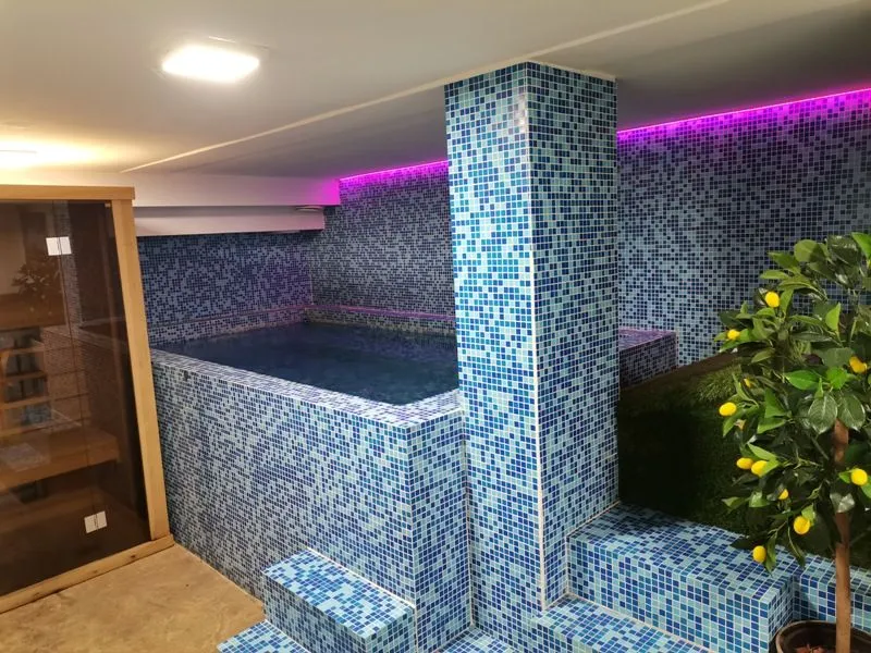 Interior of an indoor swimming pool tiled with blue mosaics featuring a sauna and decorative lighting.