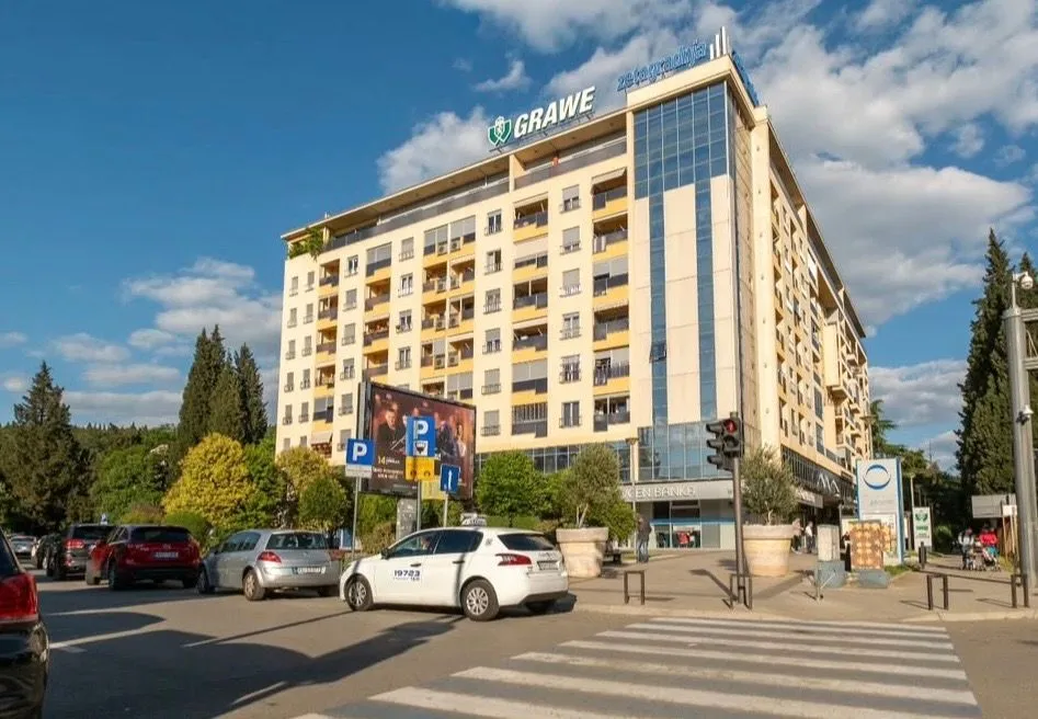 Pogled na spoljašnjost stambeno-poslovne zgrade sa parkingom i ulicom u prvom planu.