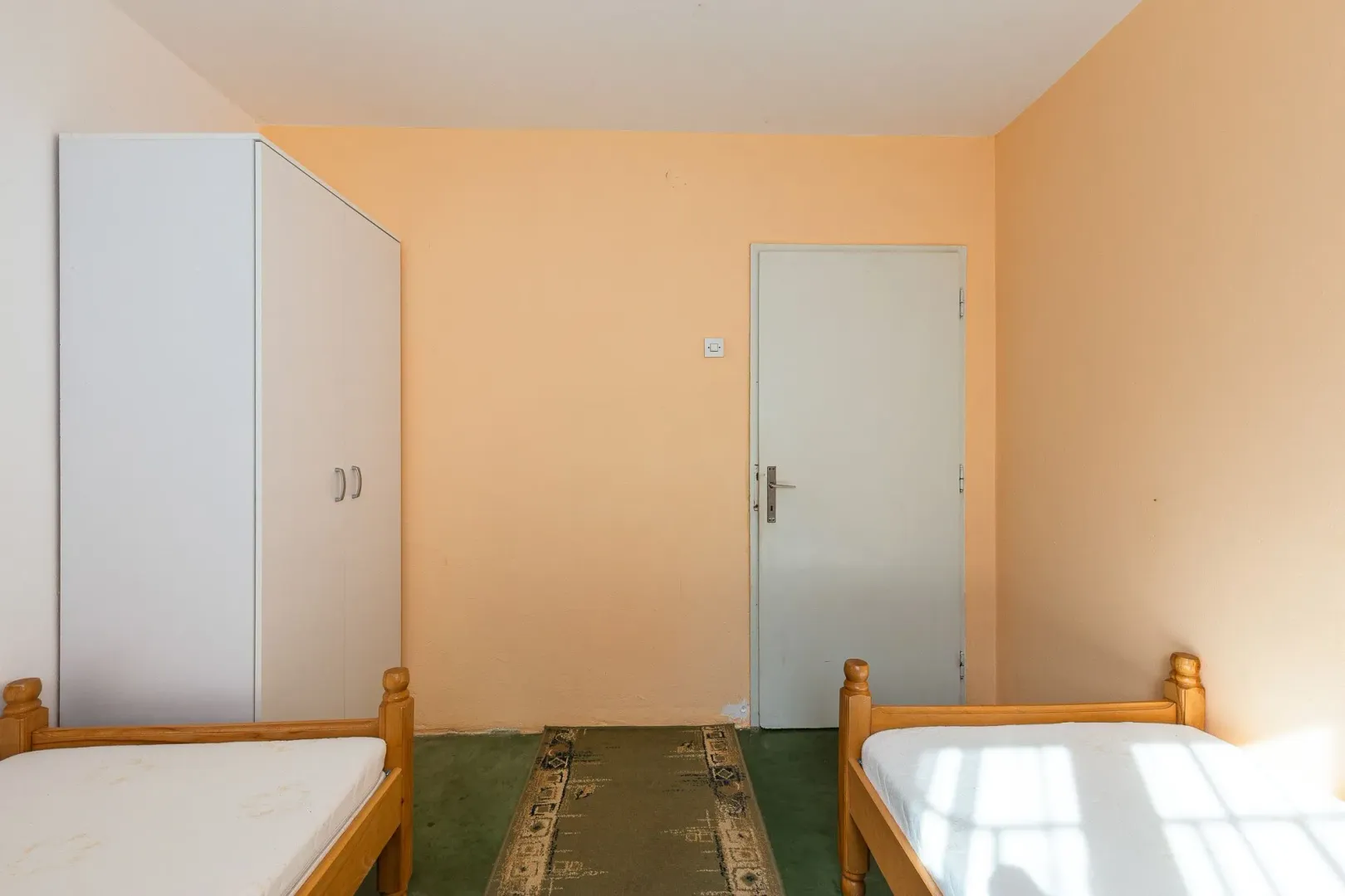 Spavaća soba sa dva drvena kreveta, bijelim garderoberom i vratima.