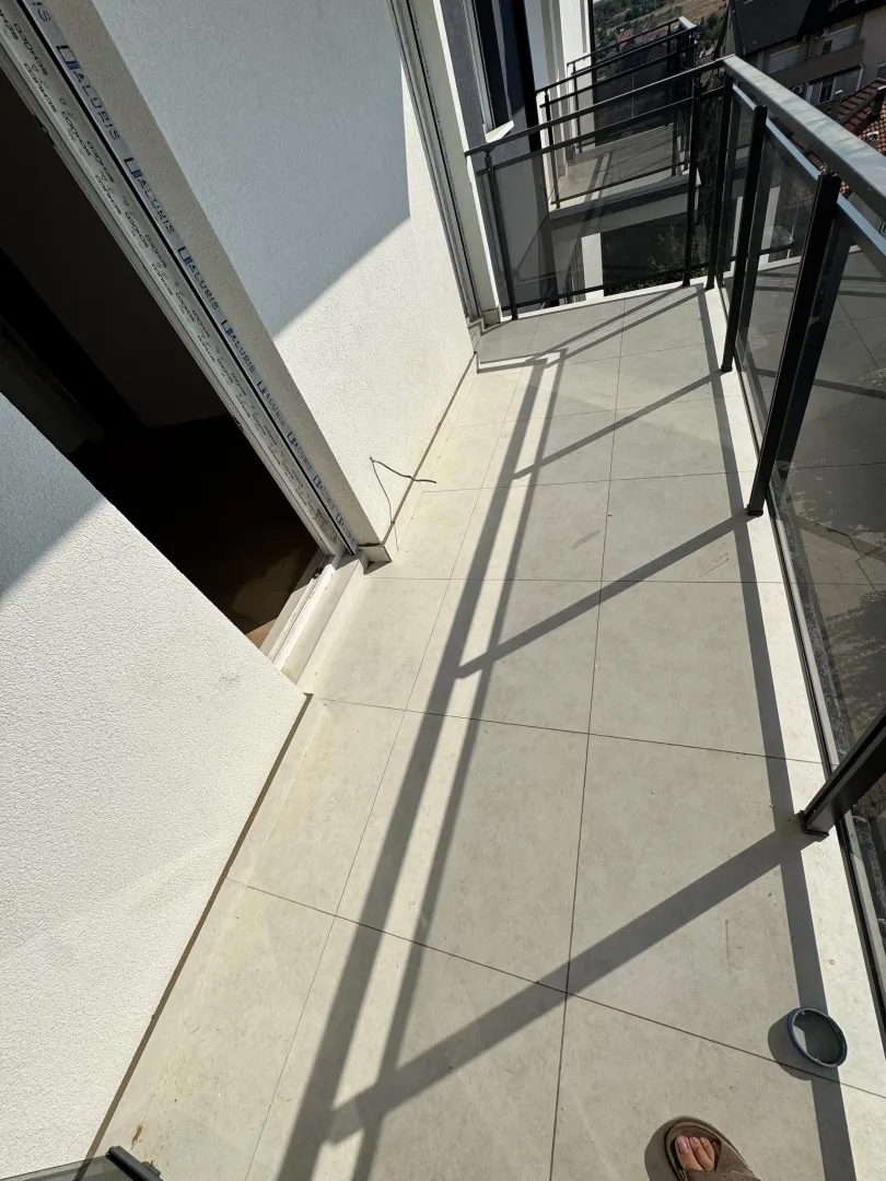Prazan prostrani balkon sa keramičkim pločicama i ogradom od tamnog metala.
