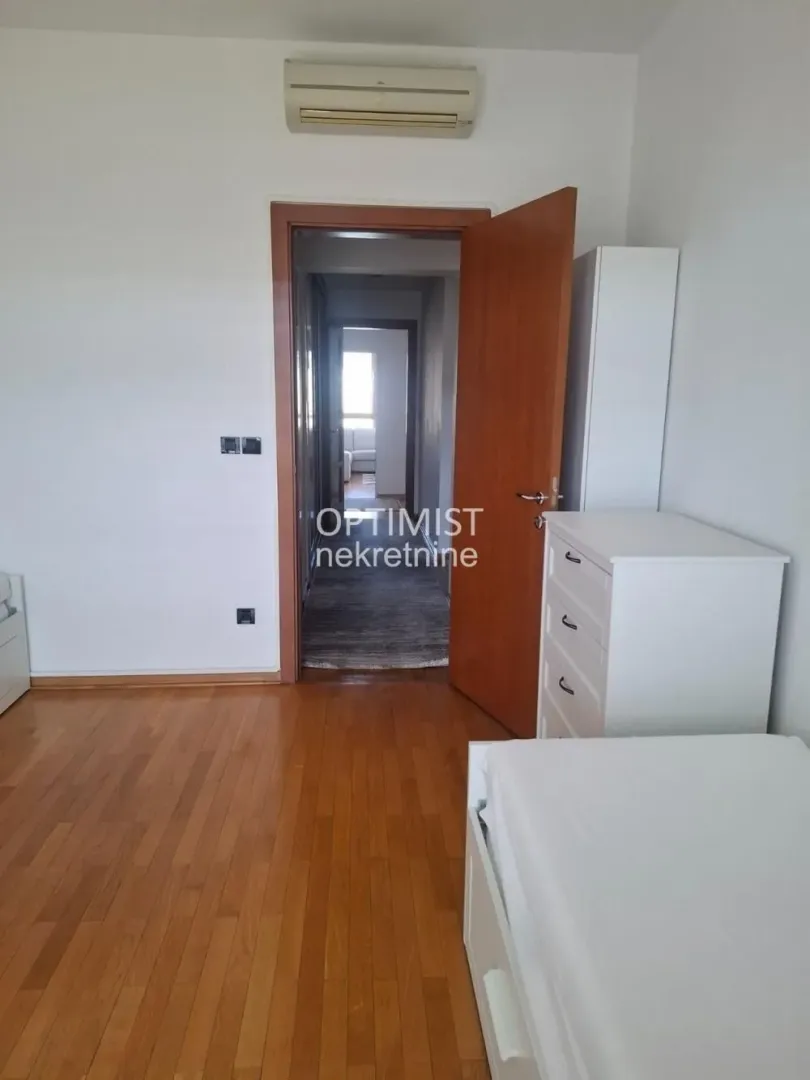 Pogled na deo spavaće sobe sa krevetom, belom komodom i otvorenim vratima.