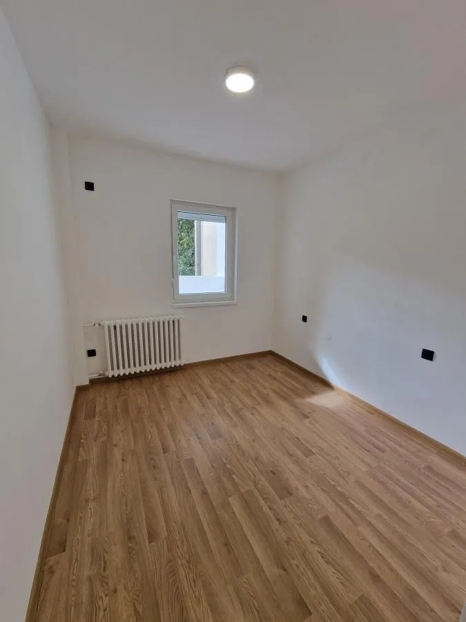 Prazna soba sa belim zidovima, drvenim podom, prozorom i radijatorom.