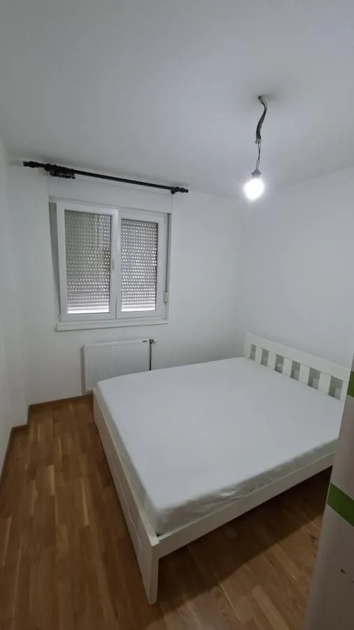 Manja spavaća soba sa bračnim krevetom, belim zidovima i drvenim podom.