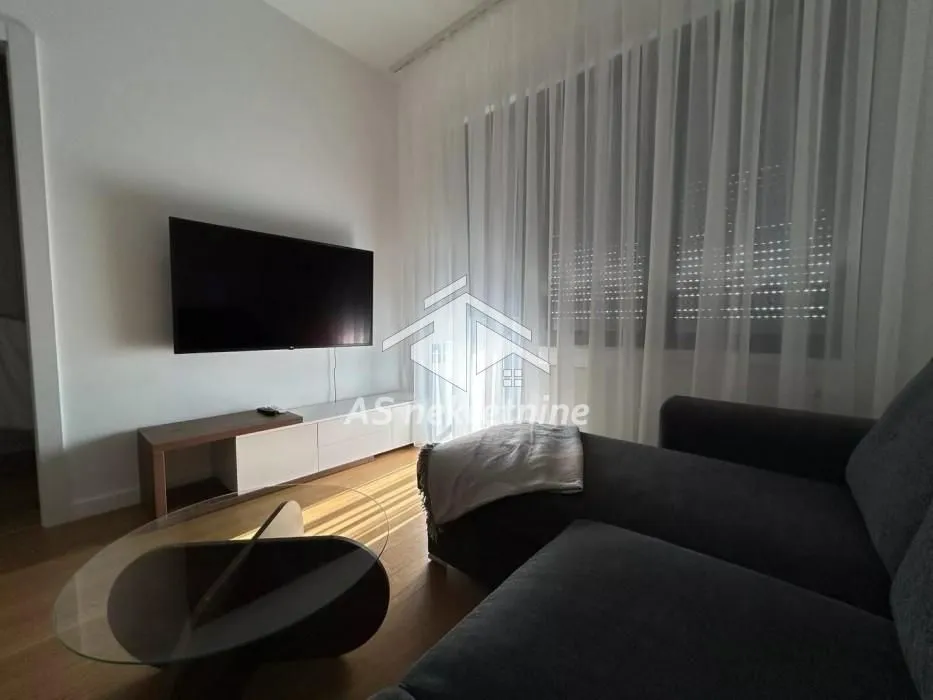 Living room with a corner sofa, wall-mounted TV, and a large window with curtains.