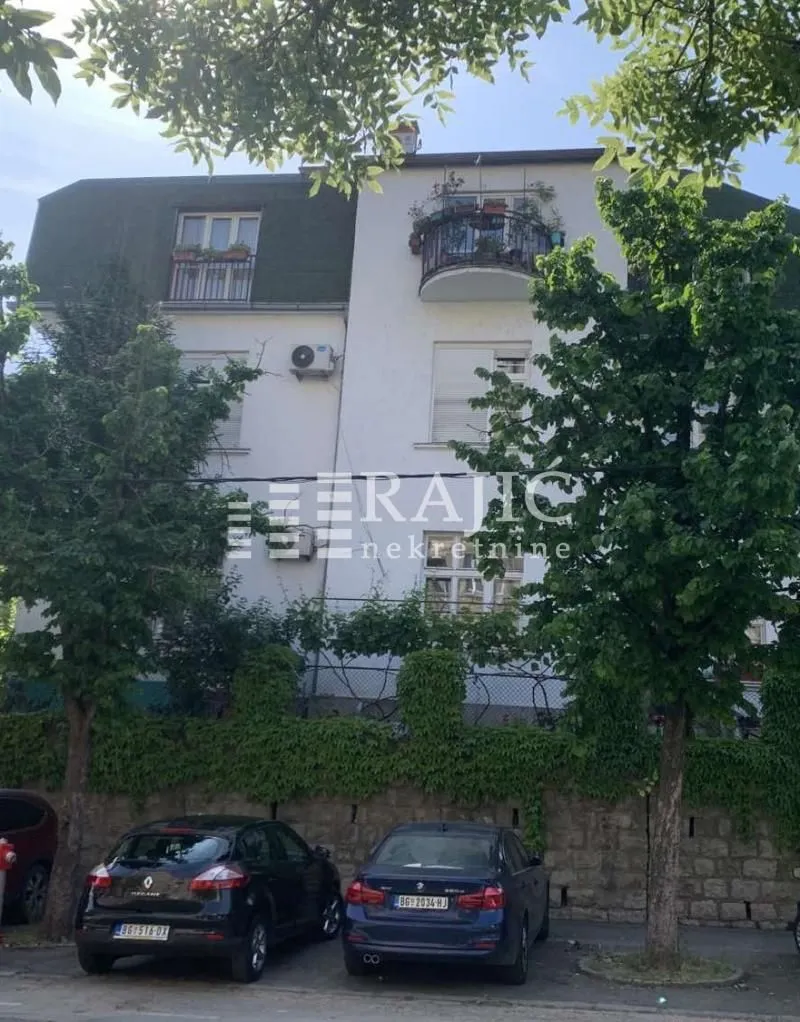 Pogled na spoljašnjost stambene zgrade sa zelenilom i parkiranim automobilima.