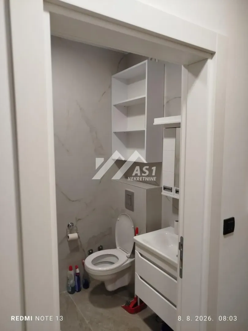 Manje kupatilo sa WC šoljom, umivaonikom i belim policama.