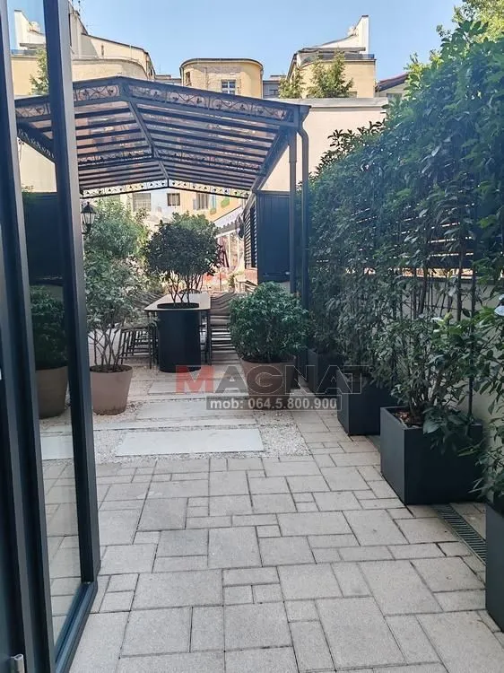 Popločano dvorište sa nadstrešnicom i uređenim zelenilom.